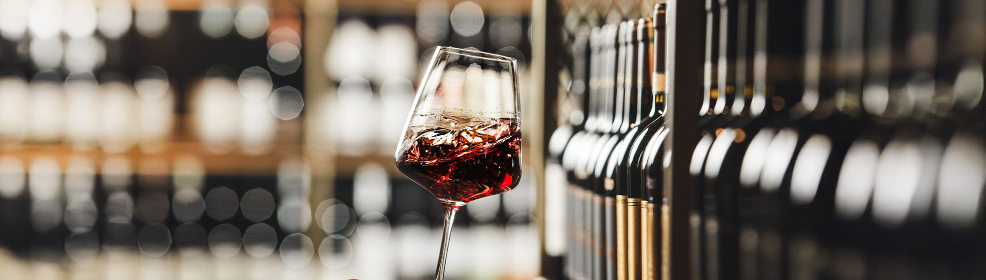 Ein Glas Rotwein steht vor einem Regal mit unscharfen Weinflaschen im Hintergrund.