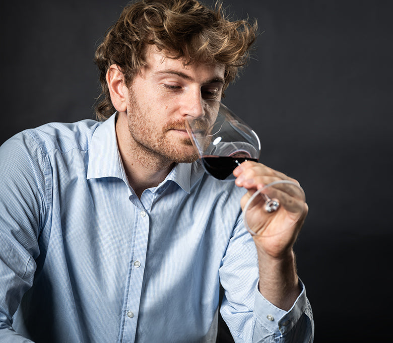Ein Mann in einem hellblauen Hemd riecht an einem Glas Rotwein vor einem dunklen Hintergrund.
