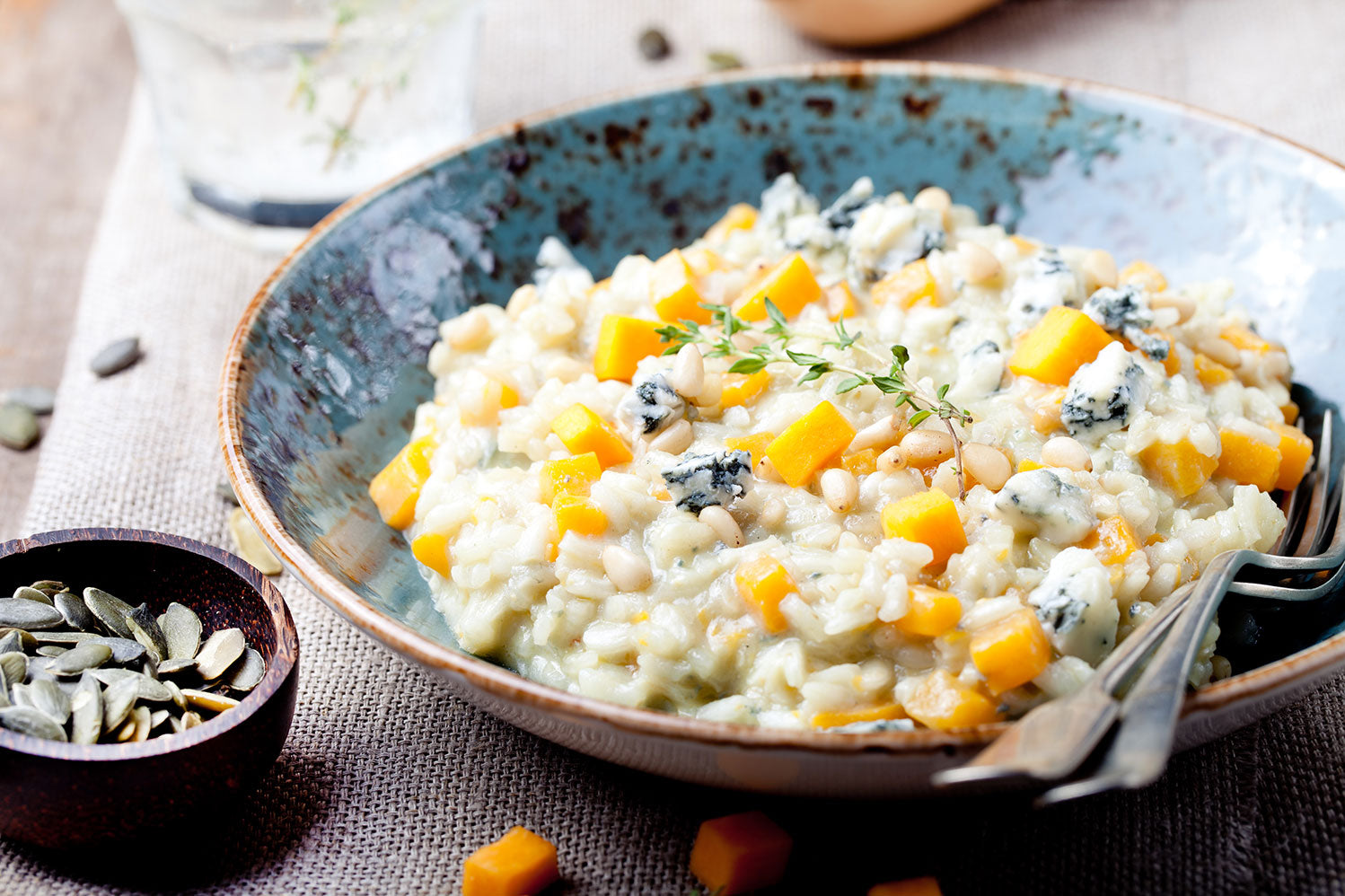 Ein Teller Risotto mit Kürbis, Blauschimmelkäse und Pinienkernen, daneben eine Schale mit Kürbiskernen auf einer Leinentischdecke.