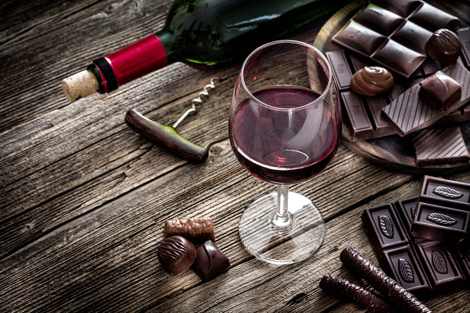Ein Glas Rotwein steht auf einem Holztisch neben einer geöffneten Flasche, Schokolade und Pralinen.