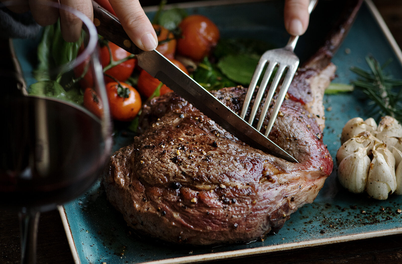 Person schneidet ein gebratenes Steak auf einem Teller mit Kirschtomaten, Knoblauch und einem Glas Rotwein daneben.