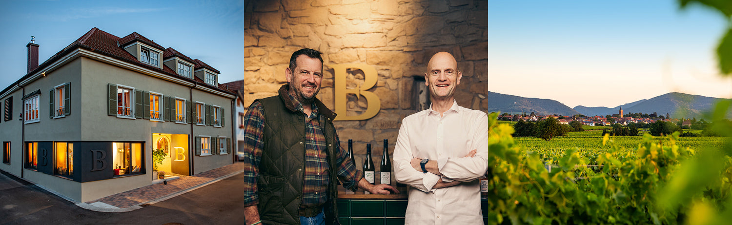 Zwei Männer lächeln vor einer Steinwand, der eine sitzt, der andere steht.