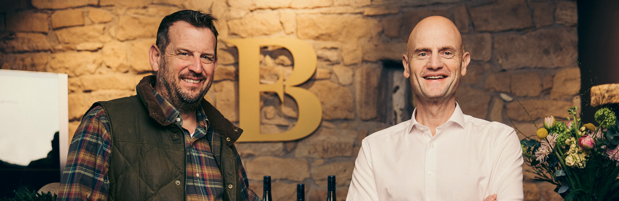 Zwei Männer lächeln vor einer Steinwand, der eine sitzt, der andere steht.