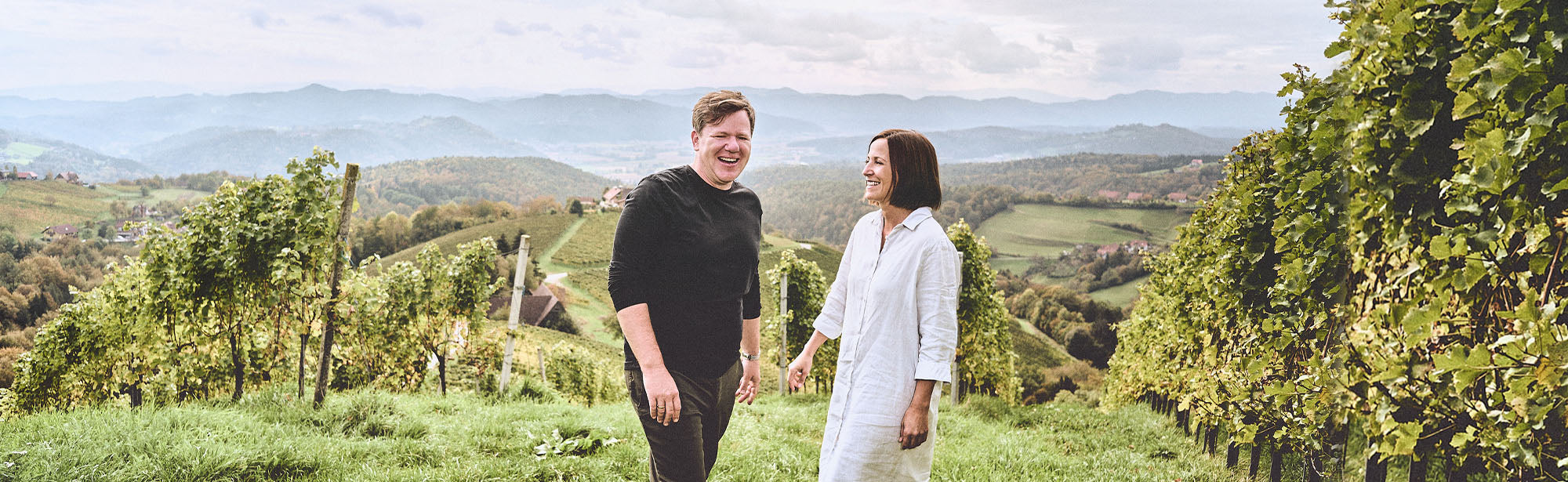 Zwei lachende Personen stehen in einem grünen Weinberg mit hügeliger Landschaft im Hintergrund.
