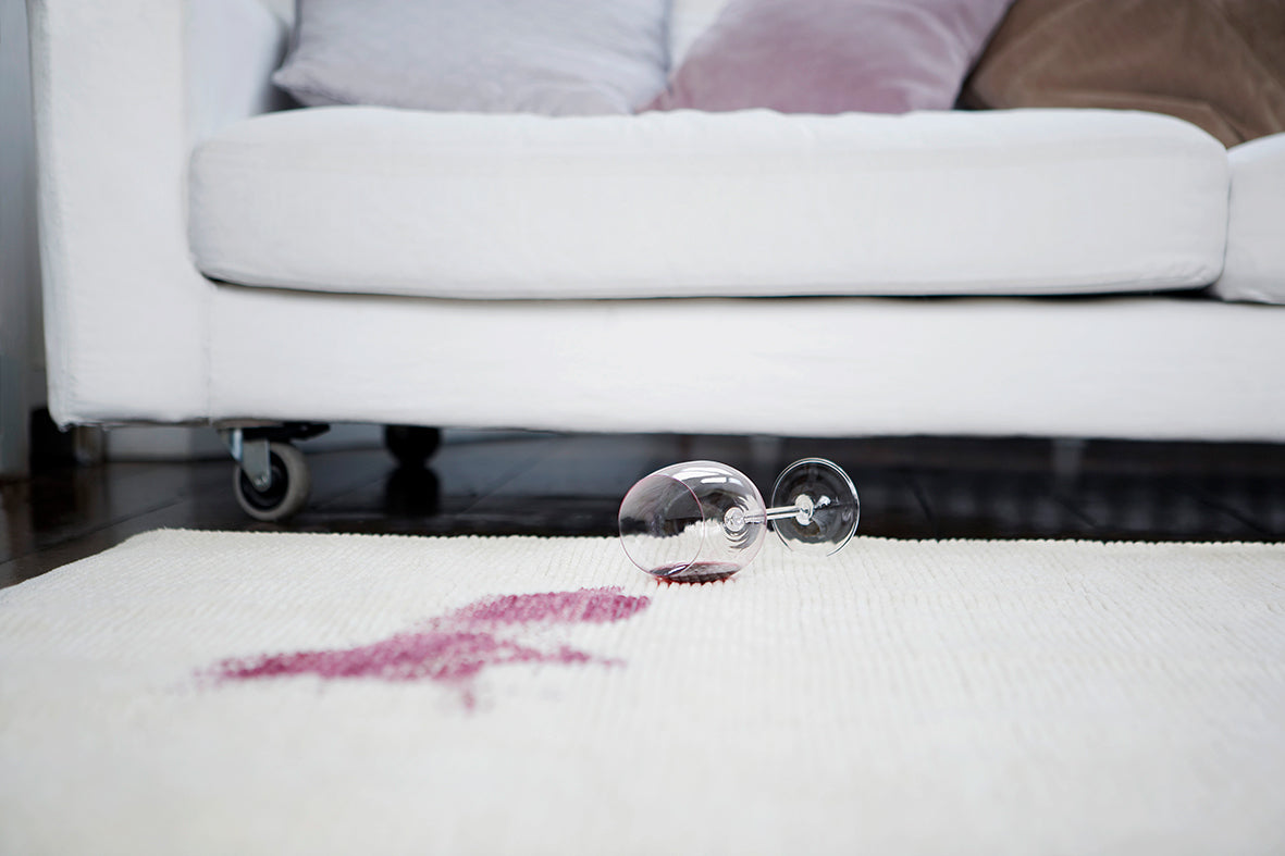 Umgekipptes Weinglas auf einem Teppich vor einem weißen Sofa, mit einem roten Fleck daneben.