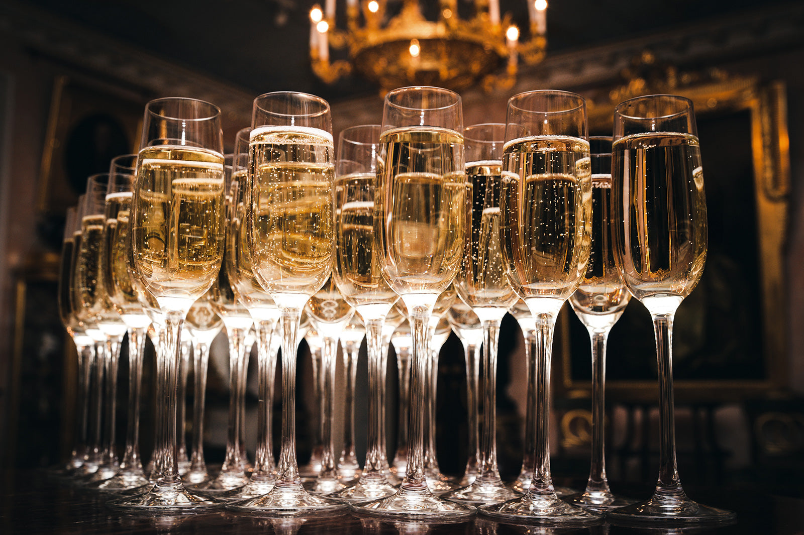 Mehrere Gläser mit Champagner gefüllt, stehend auf einem Tisch, vor einem eleganten Kronleuchter im Hintergrund.