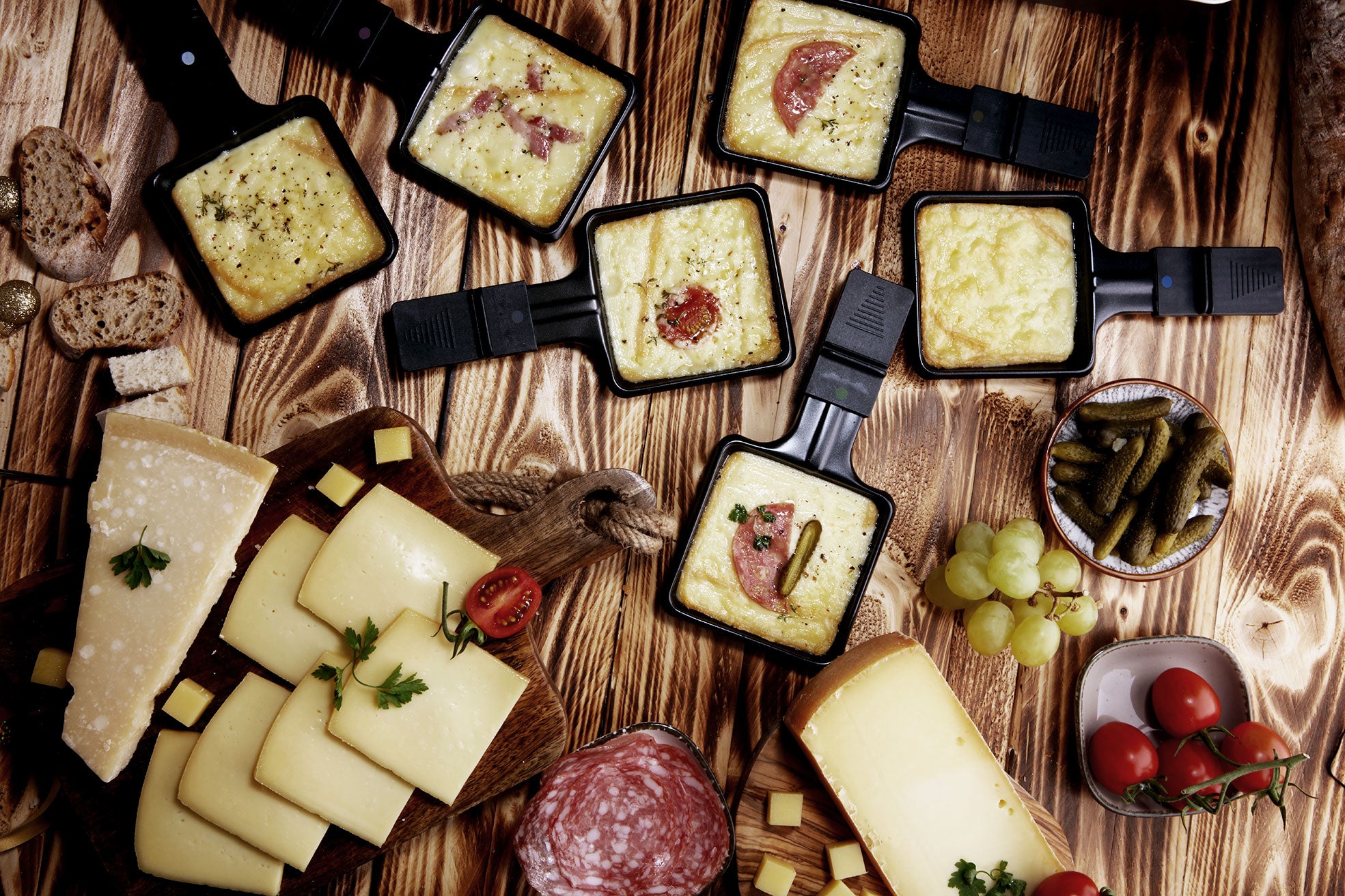 Raclette-Tablett mit flüssigem Käse und Aufschnitt, umgeben von Brotscheiben, Käse, Tomaten, Cornichons und Trauben.