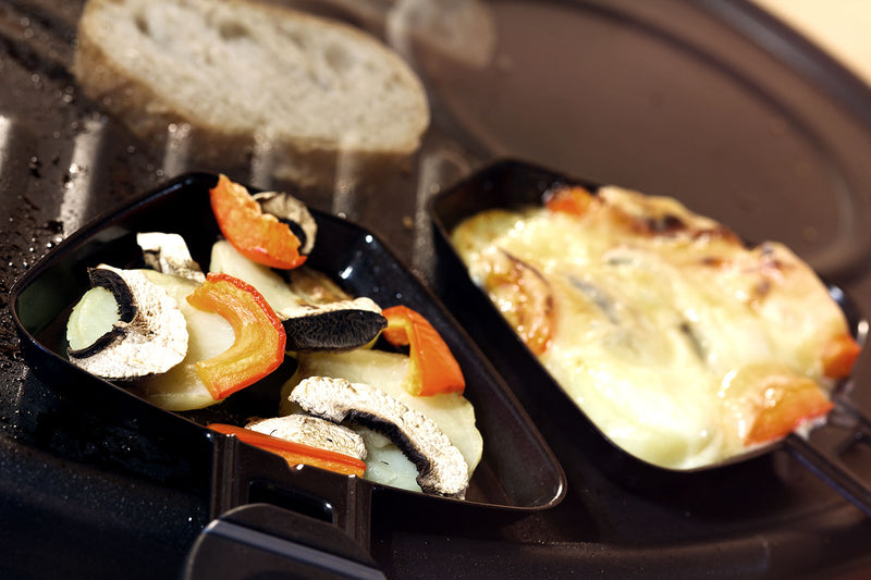 Raclette-Pfannen mit Gemüse und überbackenem Käse vor einer Scheibe Brot.