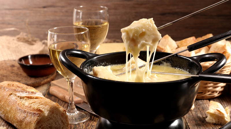 Schwarzer Fondue-Topf mit geschmolzenem Käse, Brotstücken und zwei Gläsern Weißwein auf einem Holztisch.