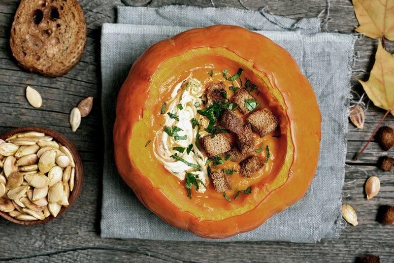Gefüllter Kürbis mit Suppe, Croutons und Petersilie, daneben geröstete Kürbiskerne und Brotscheibe auf Holztisch.