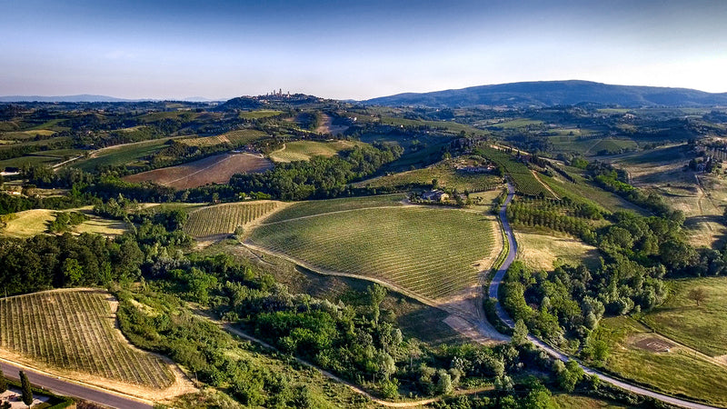 Luftaufnahme der toskanischen Landschaft mit sanften Hügeln, Weinbergen und einem fernen Dorf unter klarem Himmel.