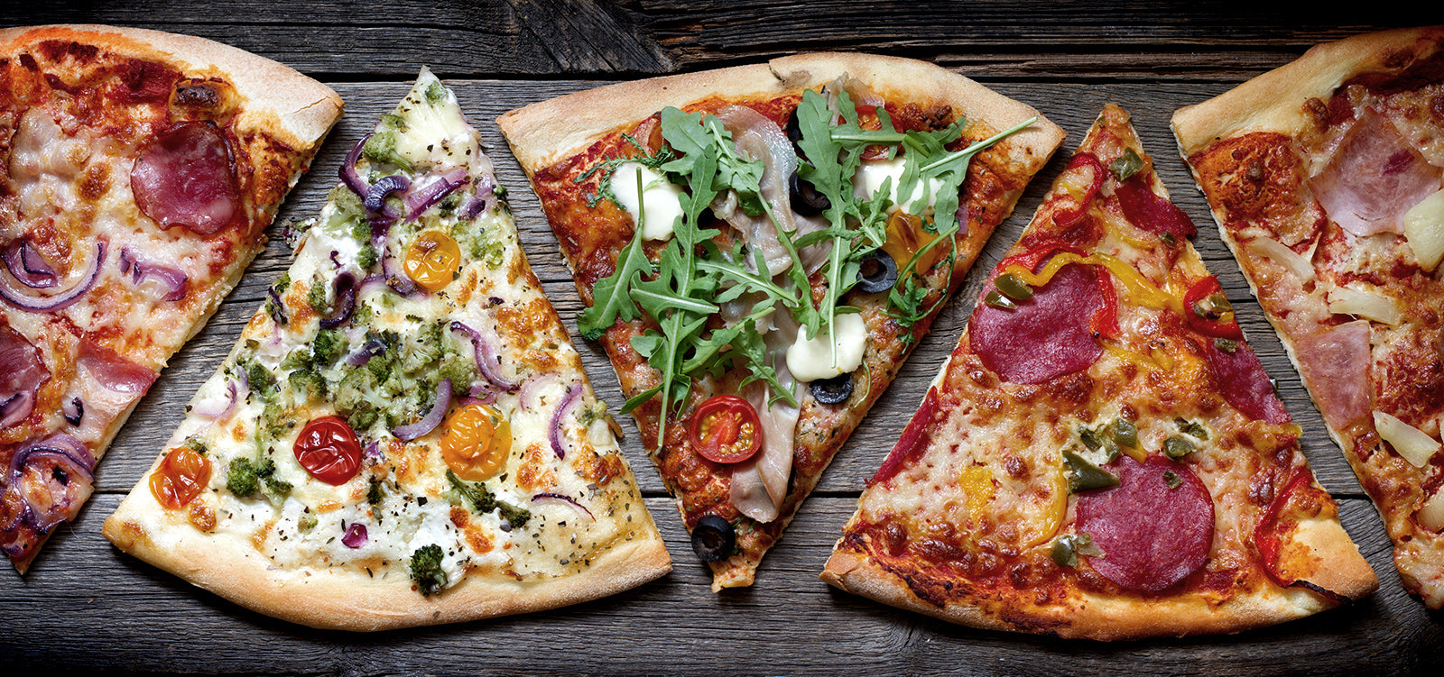 Fünf verschiedene Pizzastücke auf einem Holztisch, mit Zutaten wie Salami, Rucola, Tomaten und Peperoni.