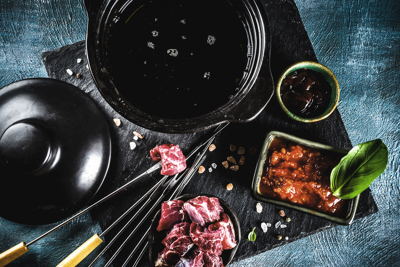 Verschiedene Schalen mit rohem Fleisch, Gewürzen und Dips auf einer Steinplatte, darauf Spieße drapiert.