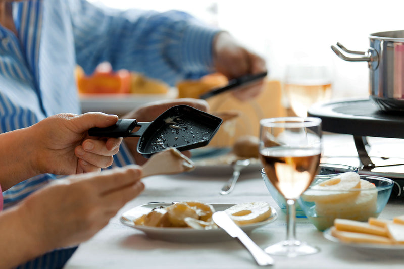 Personen bereiten Raclette an einem Tisch mit Käse, Brot und Getränken zu.