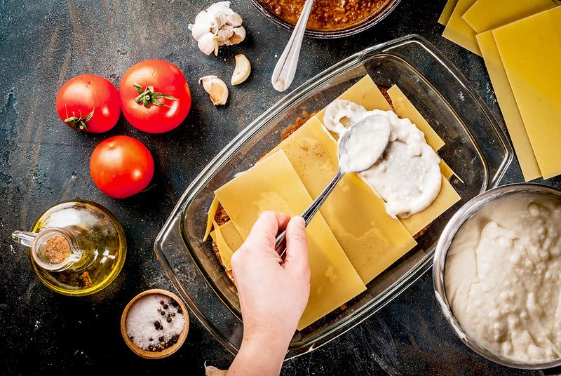 Hand verteilt Béchamelsauce auf Lasagneblättern in einer Glasform; daneben Tomaten, Knoblauch und Gewürze.